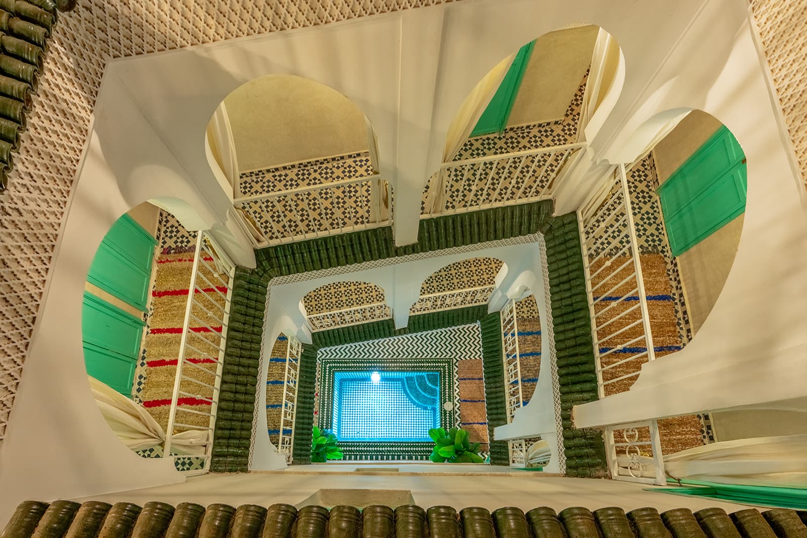 Vue plongeante depuis le toit dans le puits de lumière du riad : les arcades des deux étages, les tapis berbères, et tout au fond le bassin éclairé.