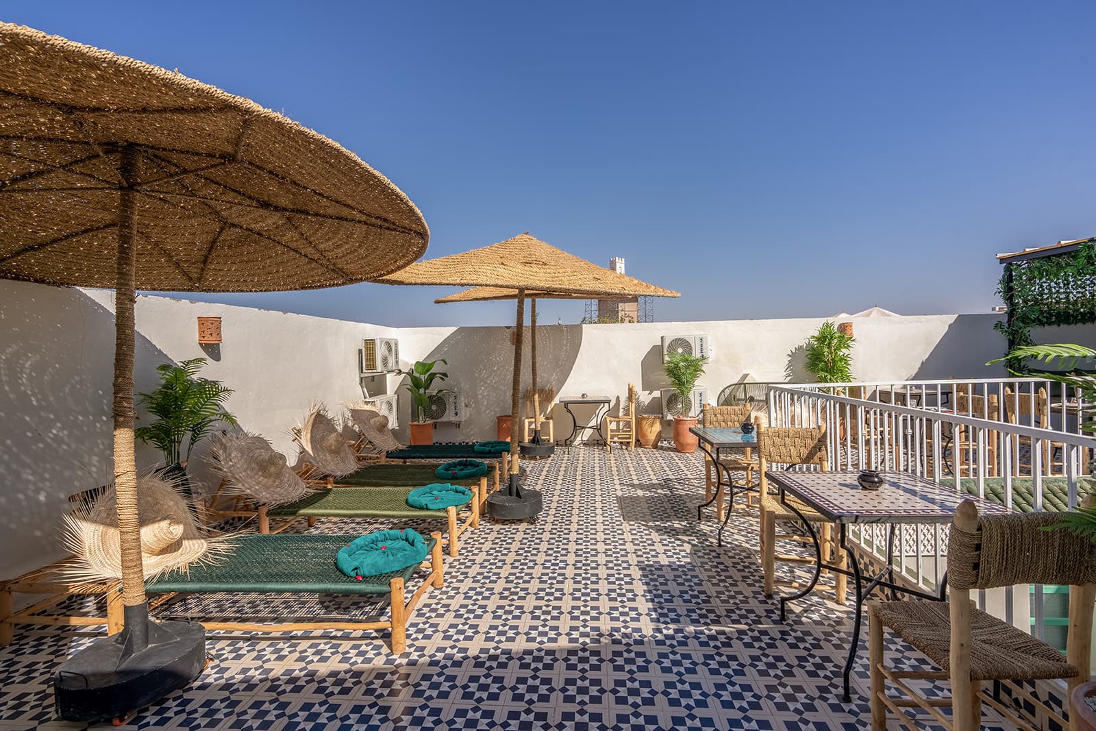Des lits de repos en bois tressé, coussins verts et chapeaux de paille posés dessus, alignés à l’ombre des parasols sur la terrasse.