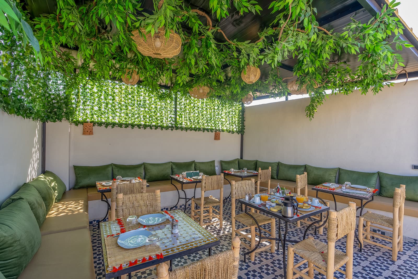Le coin repas ombragé de la terrasse, sous un feuillage suspendu et des suspensions en osier tressé, le long d'une banquette vert olive.